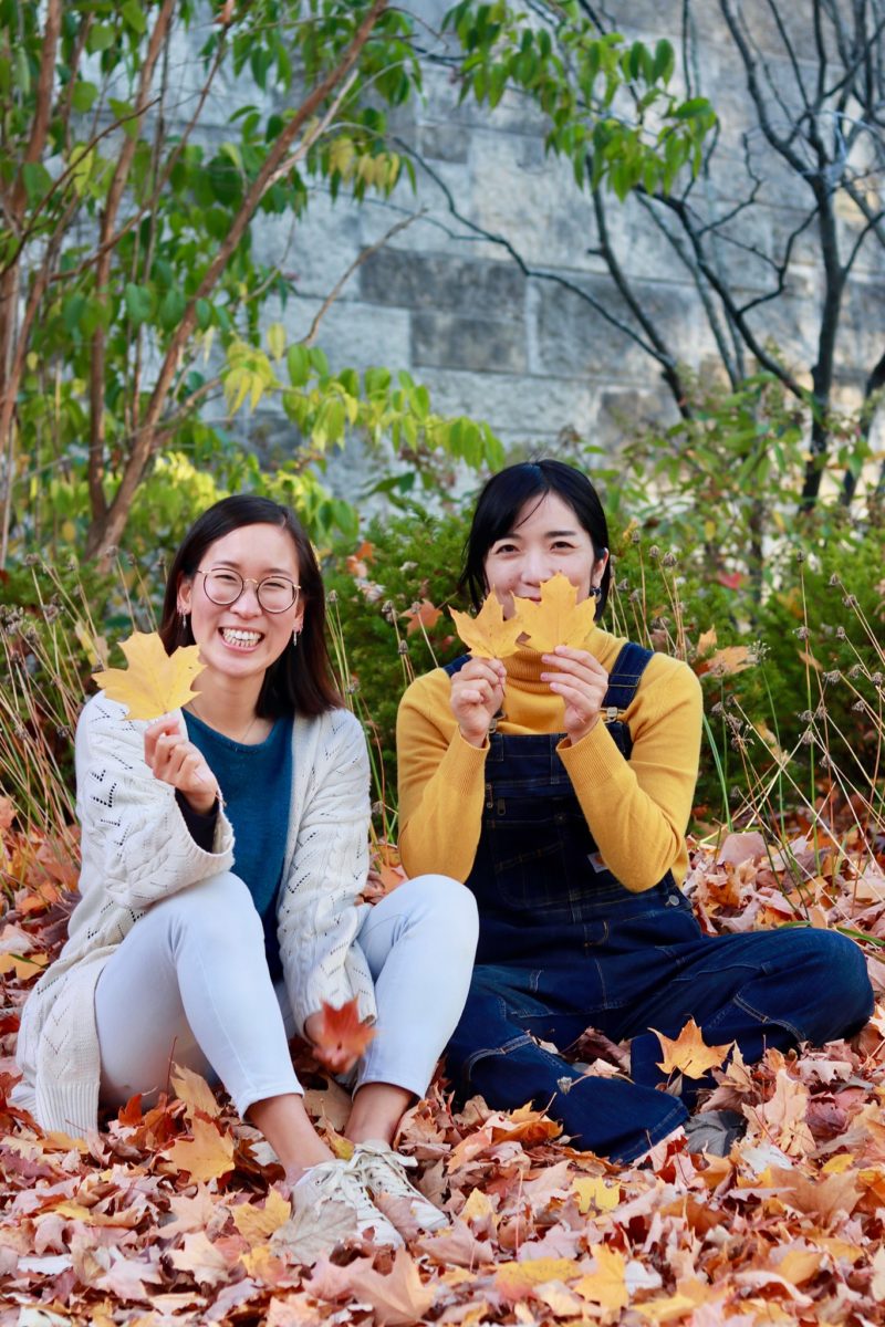 Photograph of Moe Higa and Hanna Kim sitting in fall leaves.