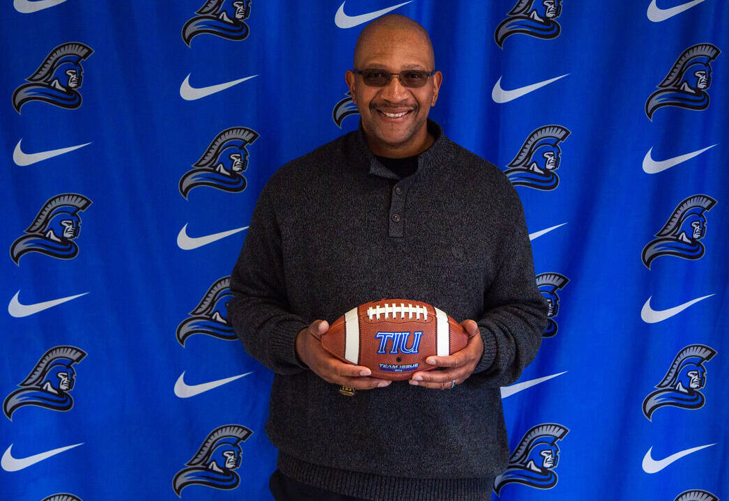 Coach Mel Mills holding the football in front of the TIU Trojans banner. 
